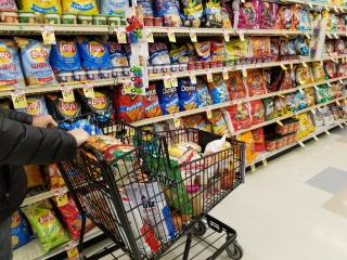A person pushes a shopping cart in the chip aisle of a grocery store.