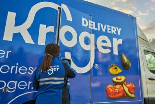 A Kroger employee stands in front of a blue truck that say "Delivery Kroger."