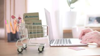 An illustration showing a person typing on a laptop next to a small shopping cart with three boxes labeled "Albertsons" inside.