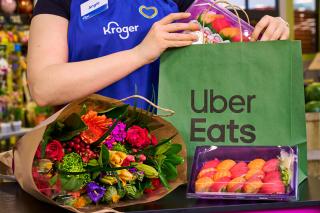 A person wearing a Kroger uniform places sushi into an Uber Eats bag. Flowers sit on a table.