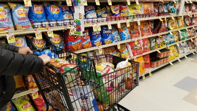 A person pushes a shopping cart in the chip aisle of a grocery store.