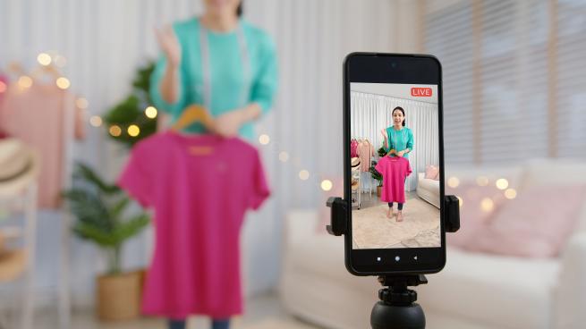 A person holds an item of clothing in front of a phone's camera that's livestreaming video.