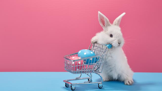A bunny pushes a small shopping cart with eggs decorated for Easter inside it.