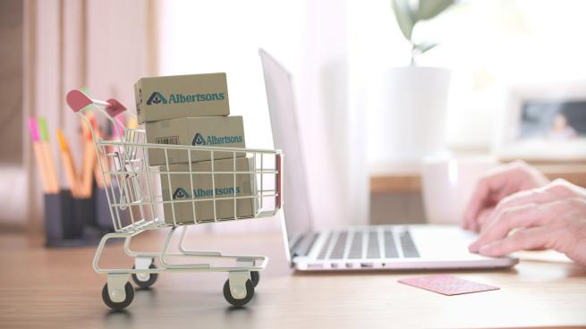 An illustration showing a person typing on a laptop next to a small shopping cart with three boxes labeled "Albertsons" inside.